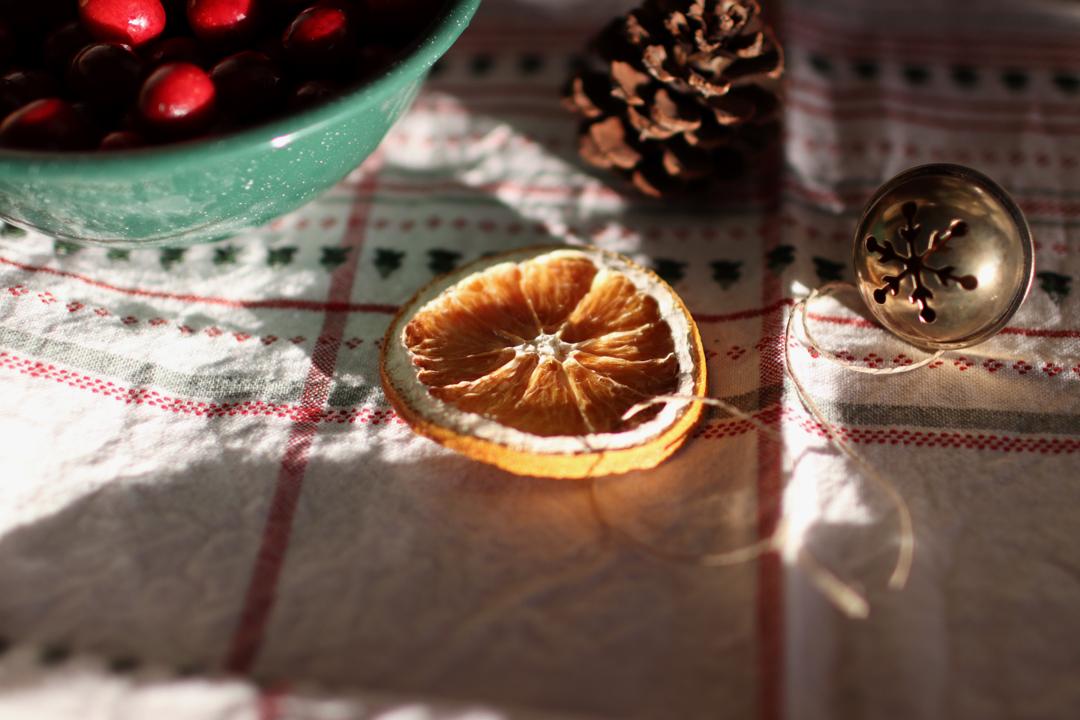 A dried orange slice, bowl of cranberries, pine cone and bell sitting on a red and white plaid towel