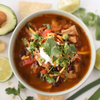 Looking down on a bowl of tortilla soup next to an avocado half, lime slices, green onions, and tortilla chips