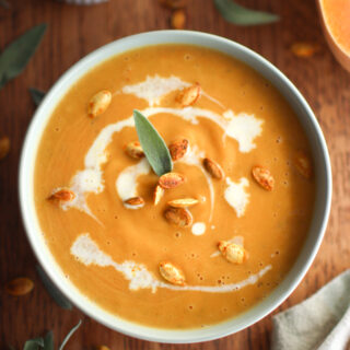 A bowl of butternut squash soup sitting on a table with a green napkin, a loaf of bread, and a sage leaf