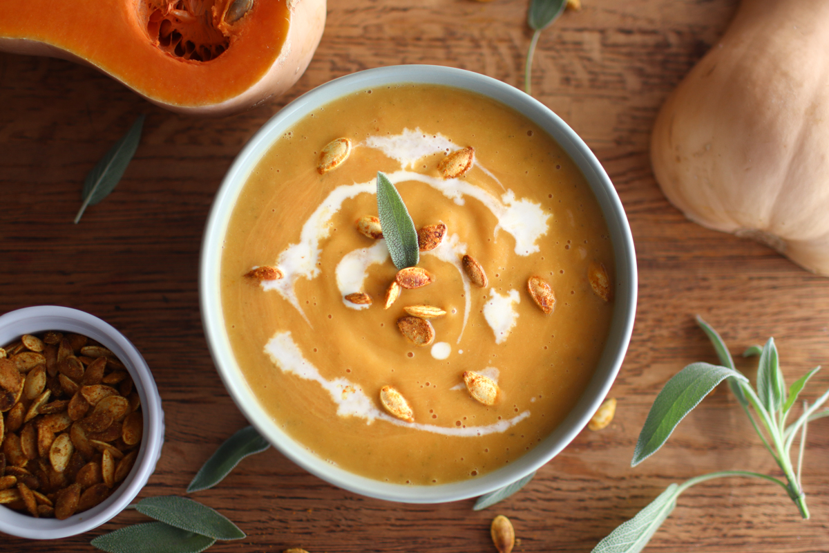 A bowl of butternut squash soup sitting on a table surrounded by a butternut squash half, a napkin, a loaf of bread, and a spoon