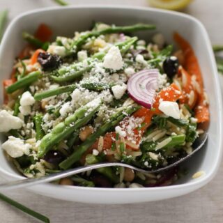 ALooking down on a bowl of lemon orzo salad filled with veggies such as peas, carrots and asparagus. There is a spoon in the bowl.