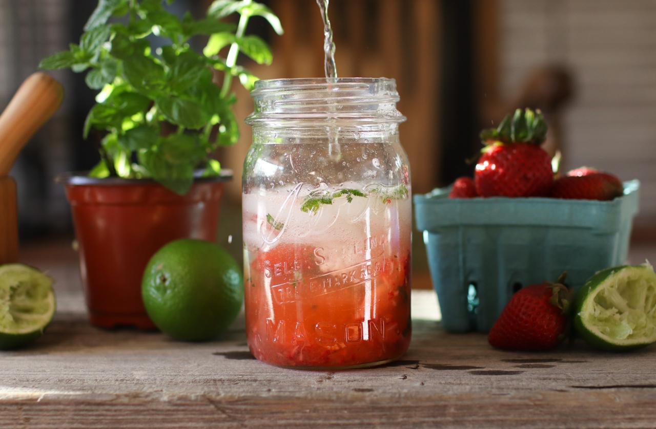 A strawberry mojito in a jar with strawberries, limes, a muddler, and mint in the background