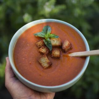 A hand holding a bowl of tomato soup with a sprig of basil and homemade croutons sitting on top.