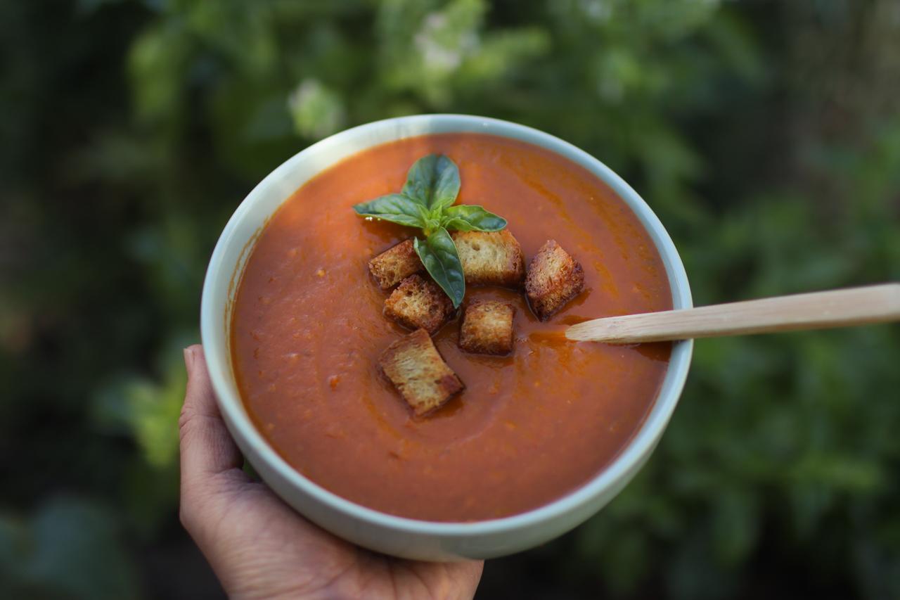 A hand holding a bowl of tomato soup with a sprig of basil and homemade croutons sitting on top.