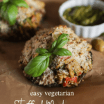 A pinterest image of a stuffed mushroom with a sprig of basil on top. In the background is another stuffed mushroom, a bowl or pesto and a green olive. The text reads "easy vegetarian stuffed mushrooms"