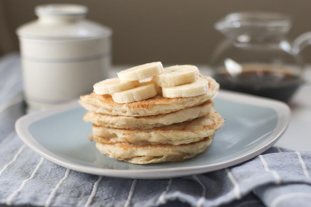 A blue plate with a stack of banana pancakes on it . Banana slices are sitting on top of the pancakes. Maple syrup and a butter bell are in the background.