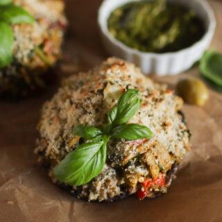 Two stuffed mushrooms with a sprig of basil on each. An olive and a bowl of pesto are in the background.