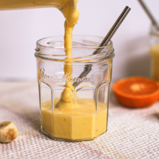 A mango smoothie being poured into a glass with banana slices, oranges, frozen mango chunks, and a second mango smoothie in the background