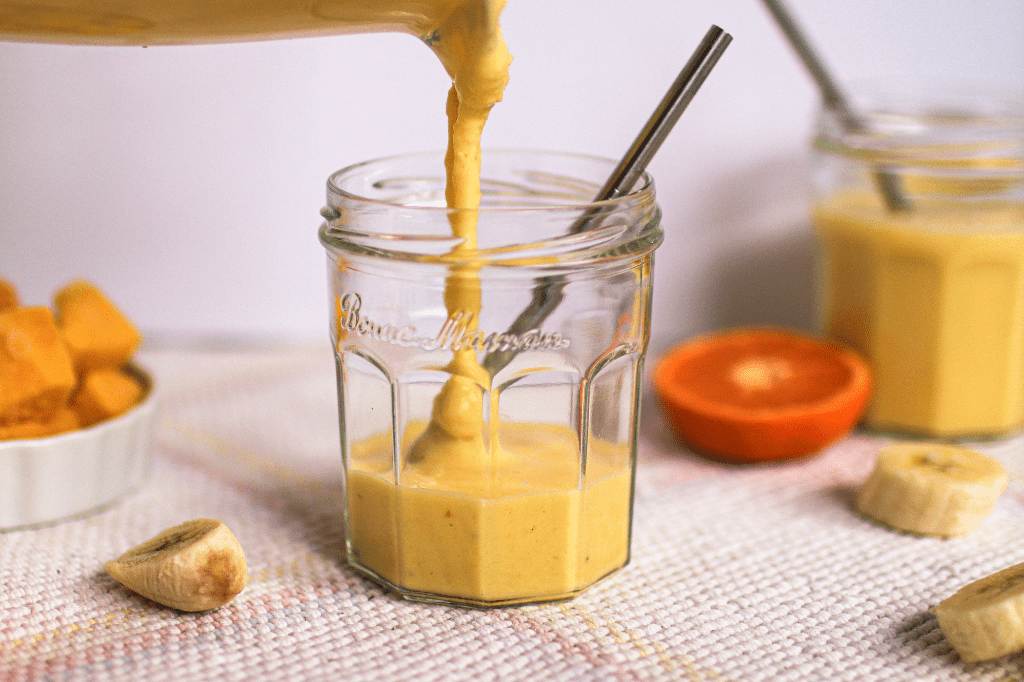 A mango smoothie being poured into a glass with banana slices, oranges, frozen mango chunks, and a second mango smoothie in the background