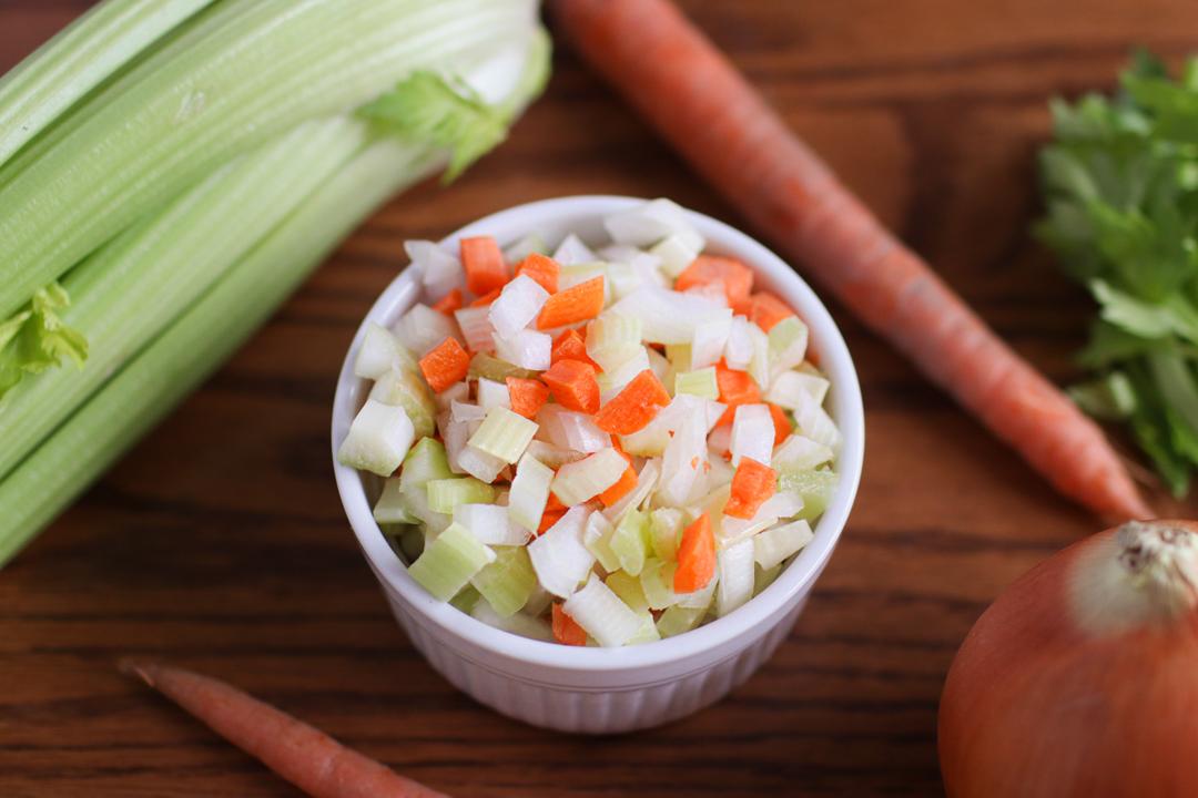 A bowl of mirepoix with celery, carrots, and an onion around it.