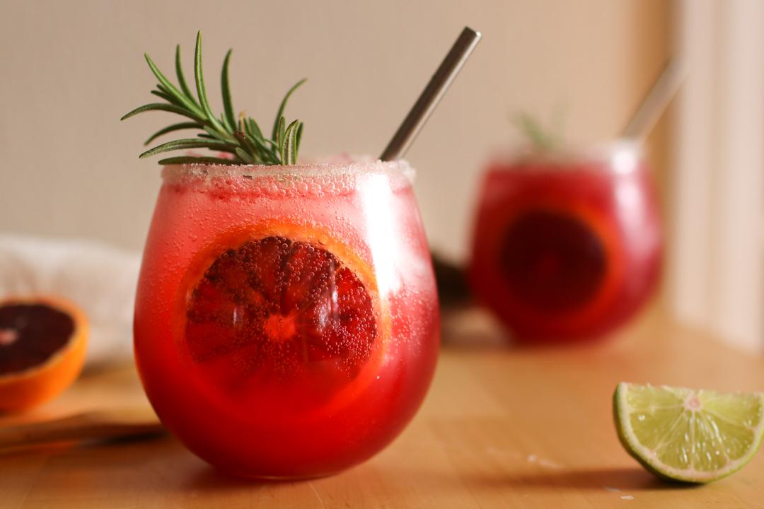 Two blood orange cocktails with a blood orange slice in the center of the glass and a sprig of rosemary sticking out the top. a lime wedge is next to them.