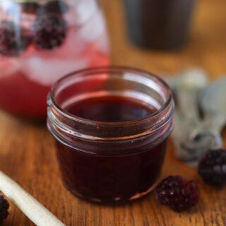 a jar of blackberry syrup with a blackberry cocktail, a honey dipper, and blackberries