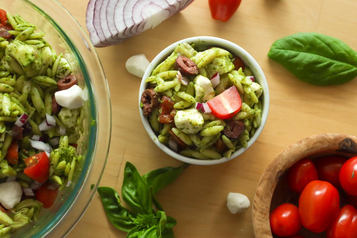a small bowl of pesto orzo salad next to a serving bowl of it. Also pictures are mozzarella pearls, cherry tomatoes, and basil leaves.