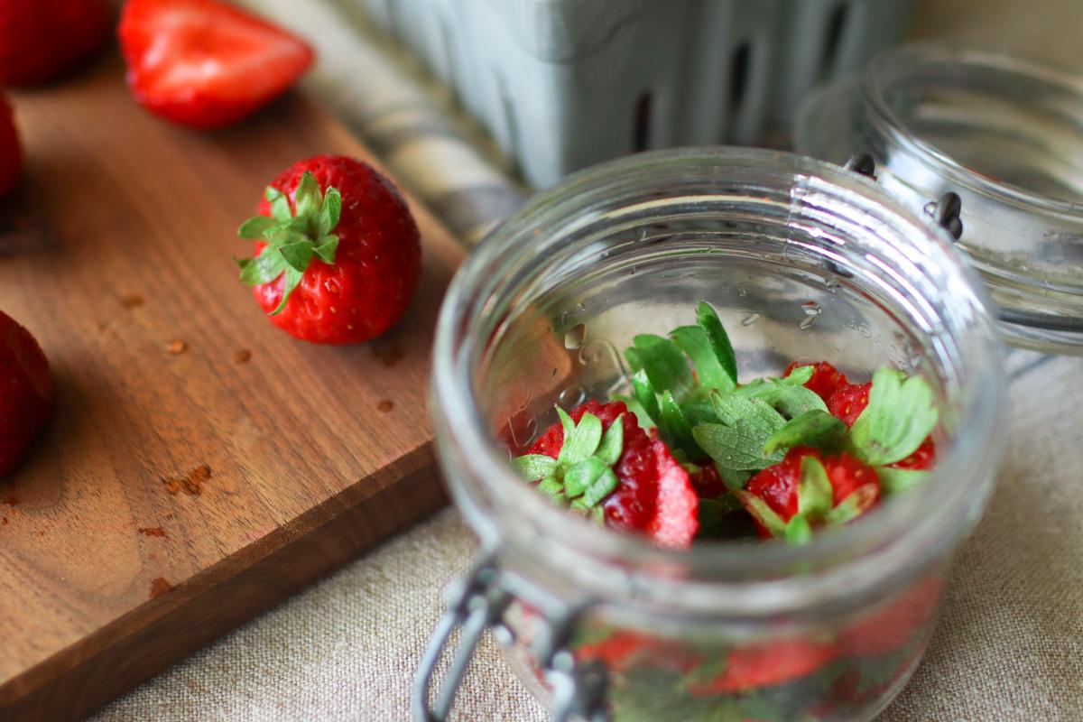 a jar of strawberry tops with a cutting board of strawberries in the background