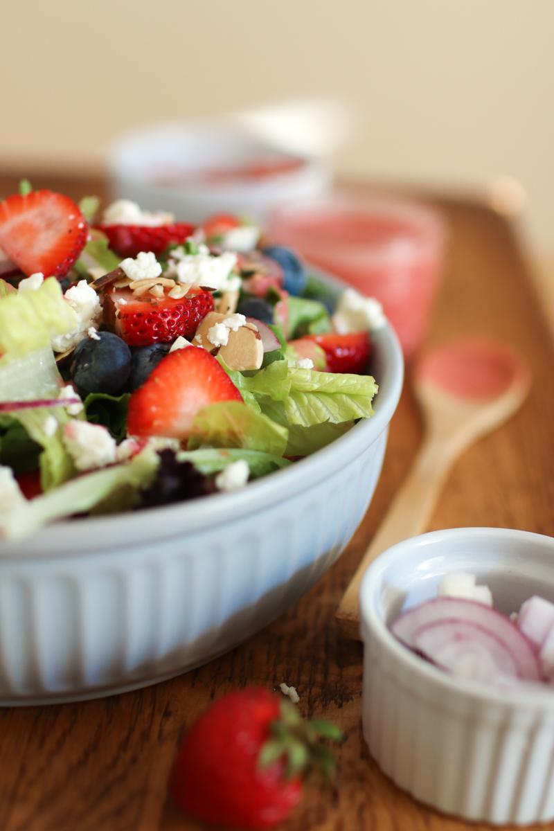 a bowl of summer berry salad with red onion slices and a strawberry next to it