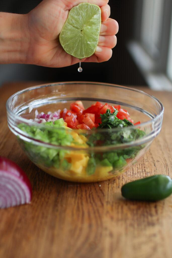 a hand squeezing half a lime onto a bowl of mango pico de gallo