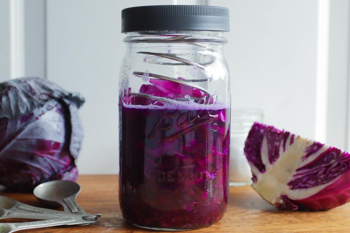 a jar of sauerkraut with measuring spoons, a cabbage head, and a chunk of cabbage