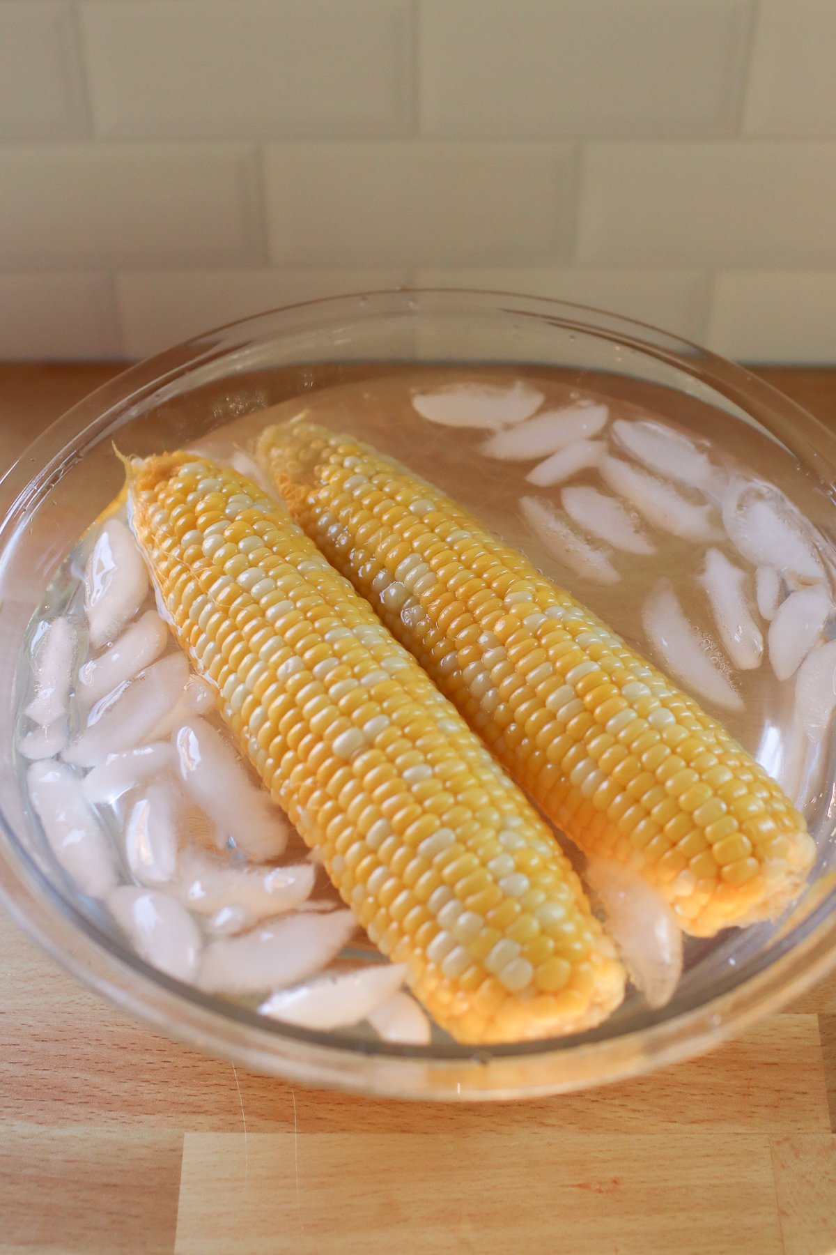 Corn on the cob floating in a glass bowl of ice water.