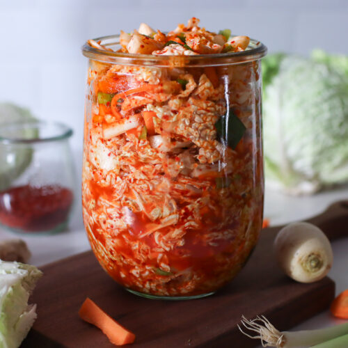A jar of kimchi sauerkraut on a wooden board with fresh veggies surrounding it.