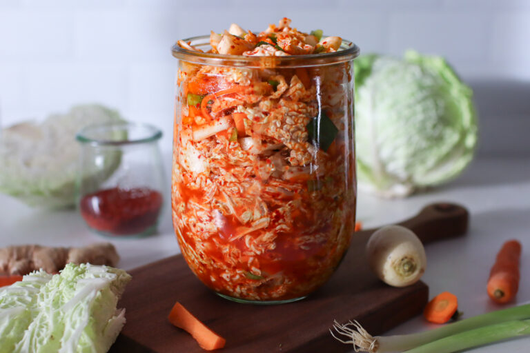 A jar of kimchi sauerkraut on a wooden board with fresh veggies surrounding it.