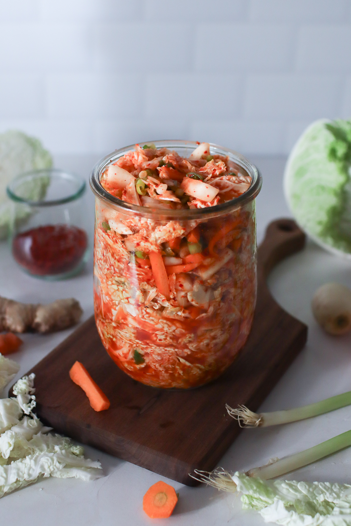 A jar of kimchi on a wooden board with kimchi ingredients sitting around it. 